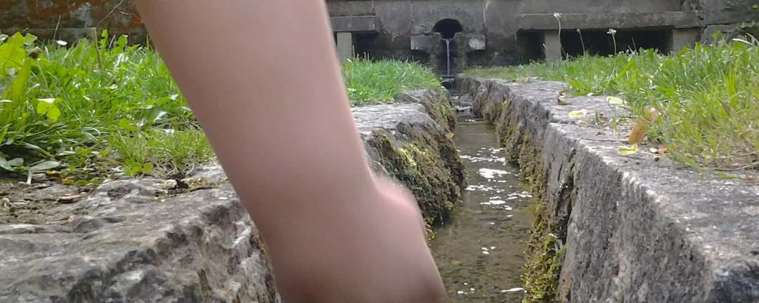 Hand im klaren Wasser eines Bachlaufs in einer alten Steinrinne – Sinnbild für Fließen und Loslassen.